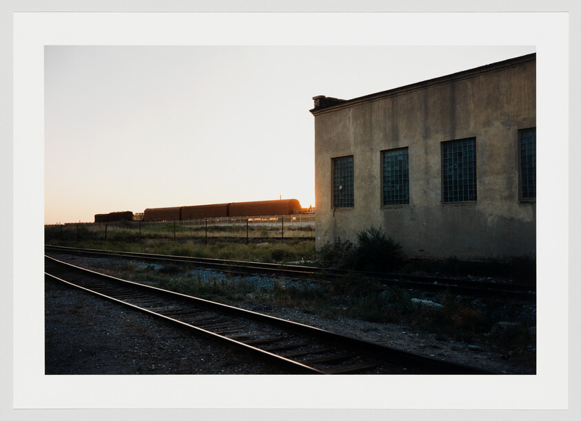 Railway tracks curve past a weathered industrial building as a freight train sits near sunset.