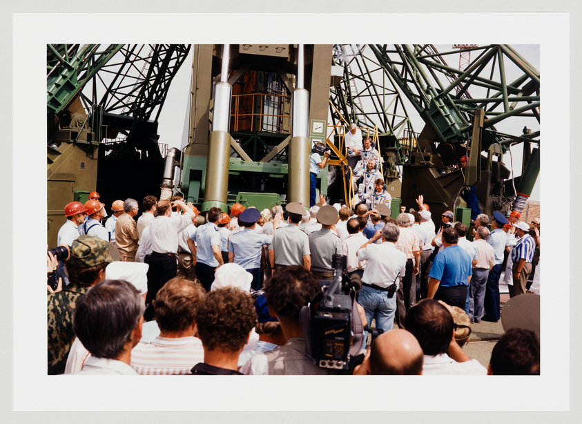 A group of people, many wearing hard hats, gather around a large piece of machinery with a complex structure, possibly at a launch site or industrial facility. Some individuals are climbing on the machinery, while others are observing from the ground. The crowd appears focused on the activity around the machinery, with some people pointing and looking upwards. A camera operator is visible in the foreground, capturing the event.