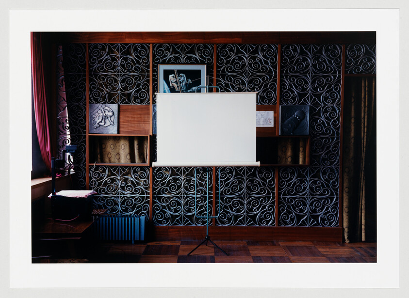 A blank white projection screen stands on a tripod in front of patterned shelving.