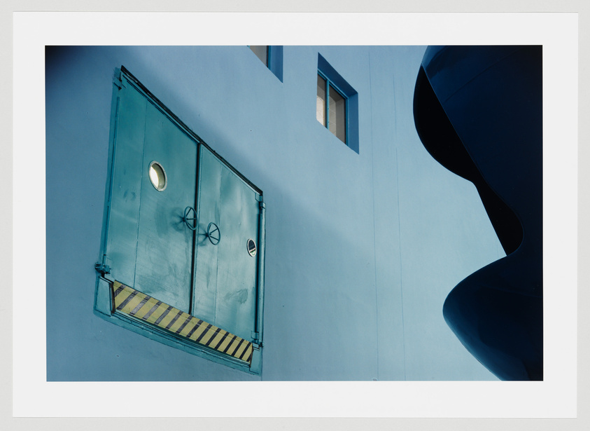 A photograph of a blue building facade with a large teal door on the left, featuring circular windows and diagonal yellow and black stripes at the bottom. On the right, there is a contrasting dark silhouette of a curvilinear structure against the light blue wall with a small rectangular window above.