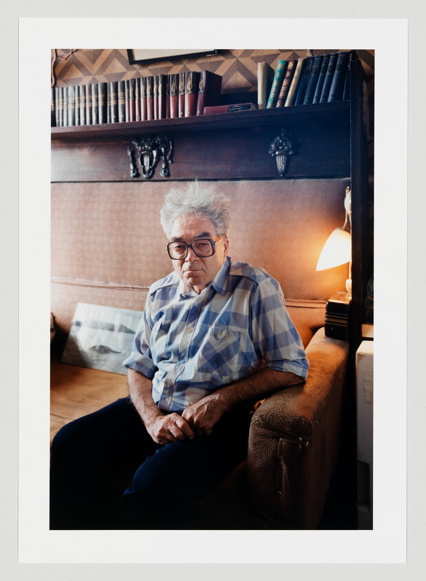Elderly man with glasses sits on a couch in a cozy room with bookshelf and lamp.