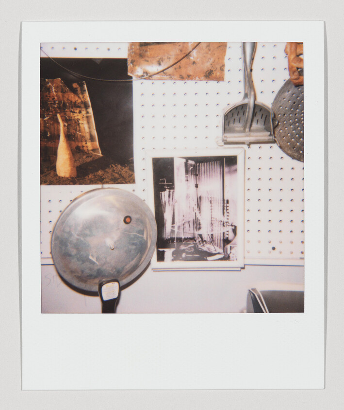 A Polaroid photo capturing a section of a wall with pegboard panels displaying various items: two photographs, one showing a natural landscape and the other an urban scene, and a couple of kitchen utensils, including a pot lid and a slotted spoon, hanging from the pegboard. The bottom edge of the photo includes a handwritten note that is partially cut off.