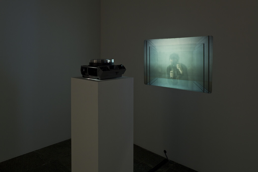 A slide projector on a pedestal displays a blurry image of a person on a nearby wall in a dark room.