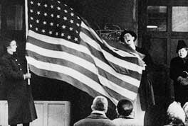 Two people hold a large American flag in front of a small group of seated onlookers indoors.