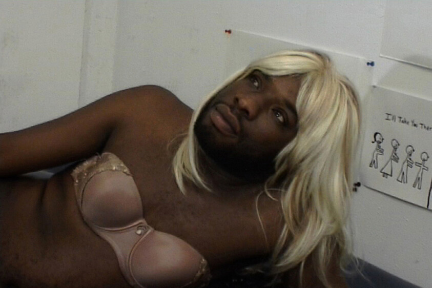 Man with a beard wearing a blonde wig and bra, looking upwards with a contemplative expression.