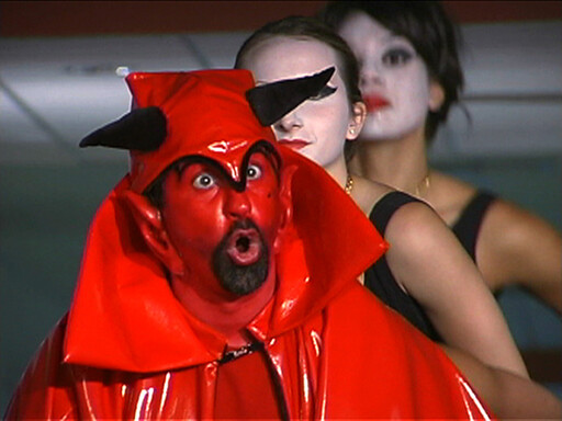 A man in a shiny red devil costume makes a surprised face while performers stand behind.