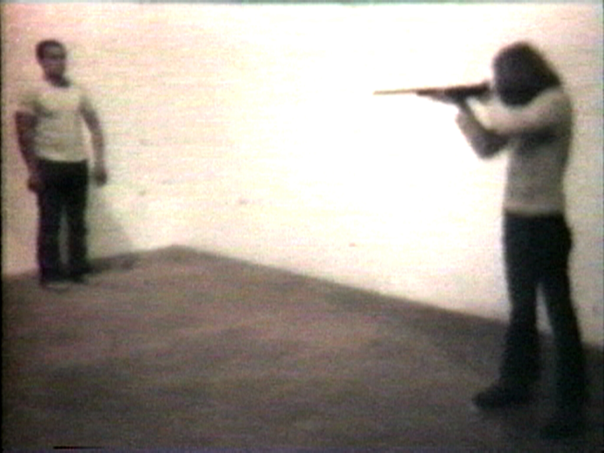 A person aims a rifle at another person standing against a blank wall in an empty room.