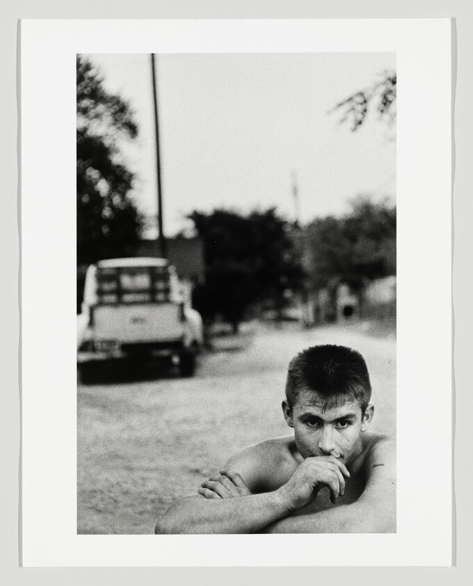 Young shirtless man resting his arms and staring at the camera on a quiet dirt road.