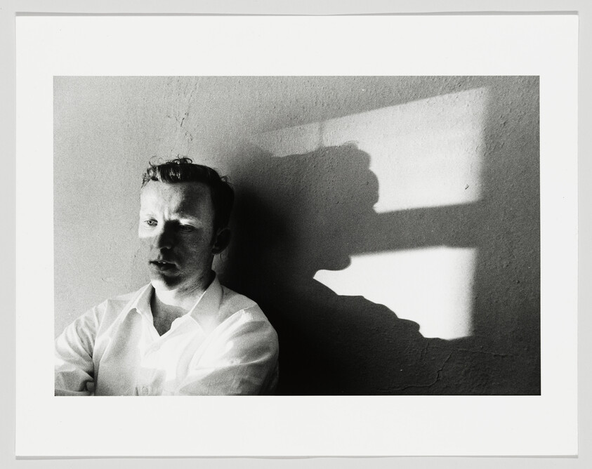 A man in a white shirt sits against a wall while a window-shaped shadow falls beside him.