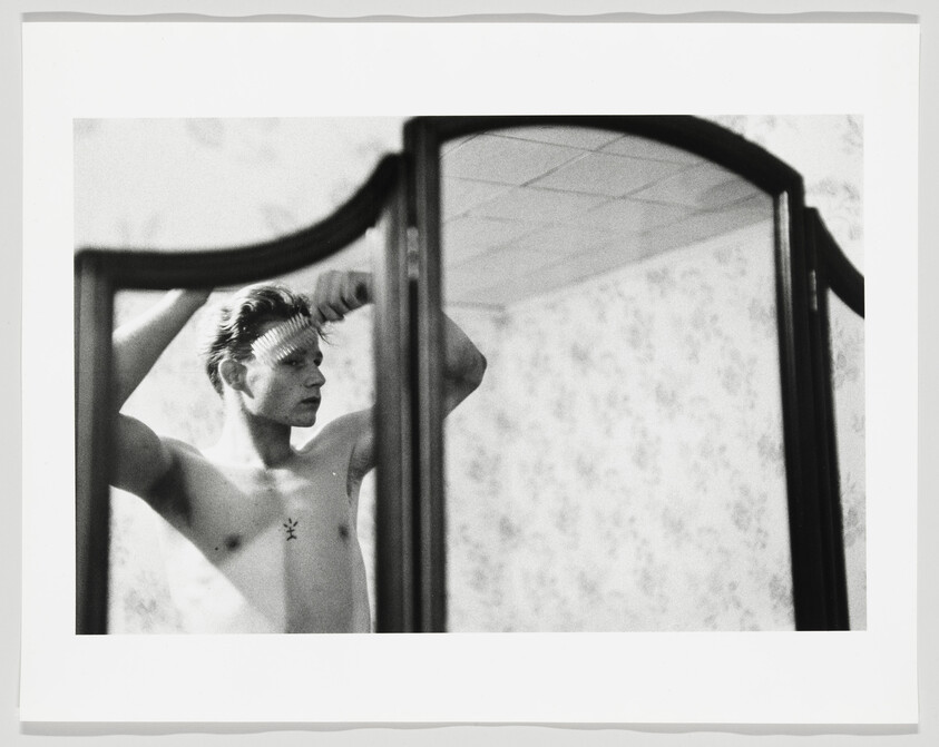 Shirtless young man combs his hair while looking at himself in a three-panel mirror.