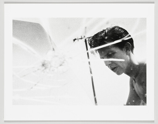 Young shirtless man leans forward while looking down through cracked glass with light streaks.