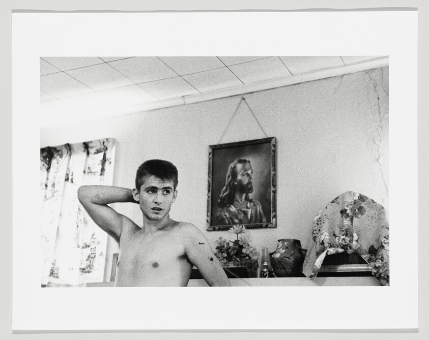 A shirtless young man with his hand behind his head stands in a room with a framed Jesus portrait.