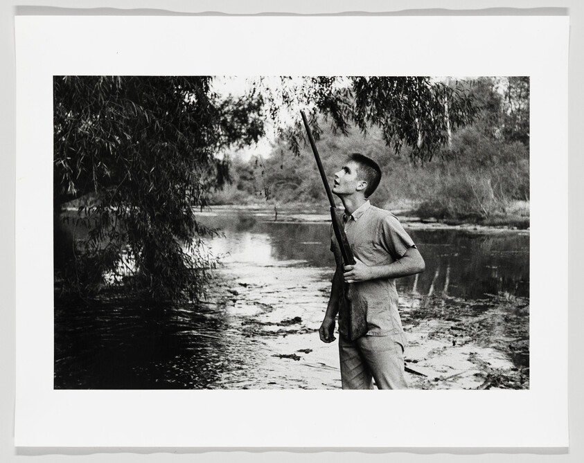 A young man stands by a river holding a rifle and looking up into a tree.