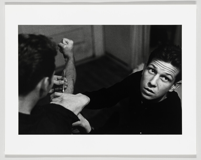 Young man holds another's arm while he injects a syringe.