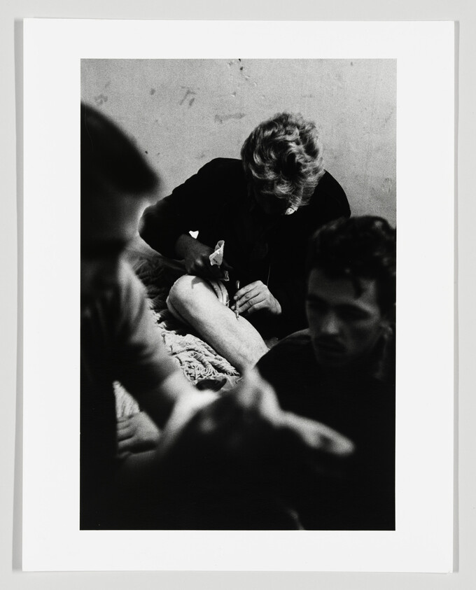 A person uses a syringe on their thigh while two others sit nearby in a dim room.