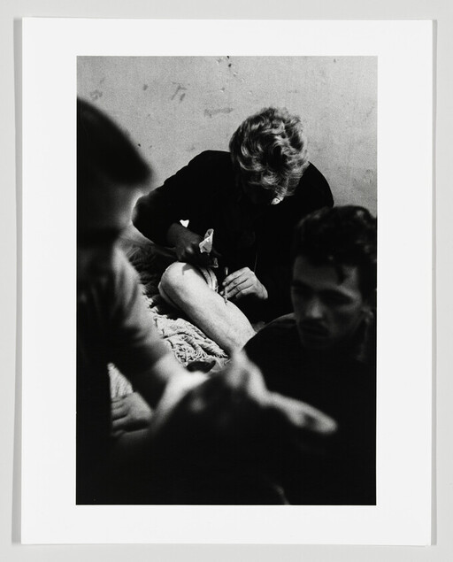 A person uses a syringe on their thigh while two others sit nearby in a dim room.