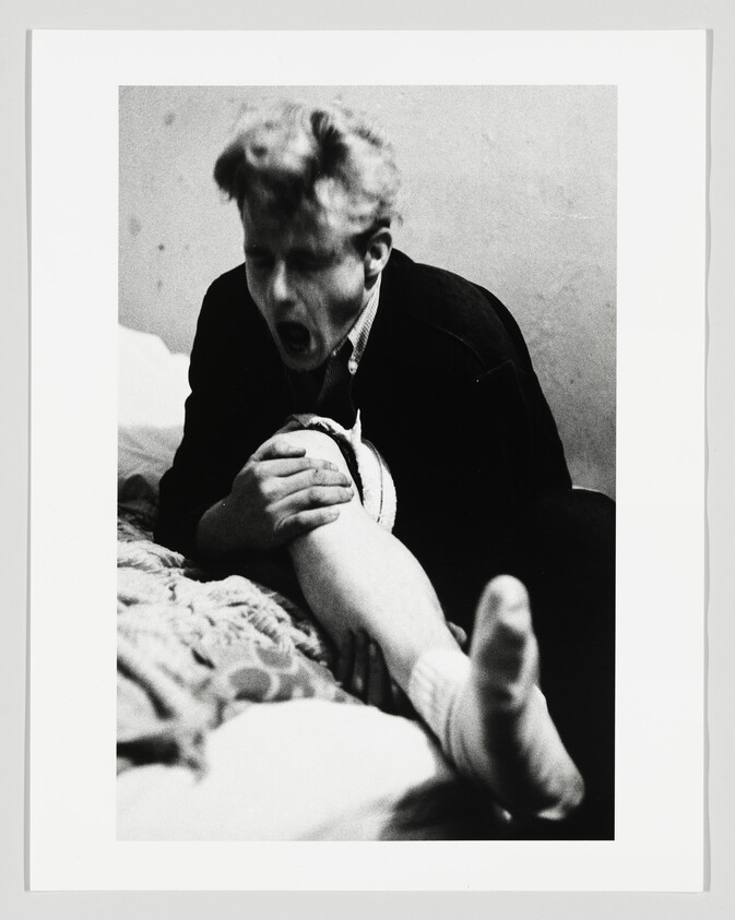 Young man grasps and appears to bite another person's knee while sitting on a bed.