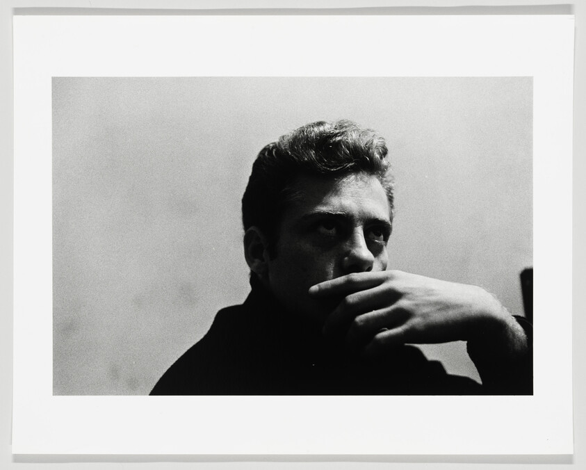 A young man sits pensive with his hand covering his mouth, staring off to the side.