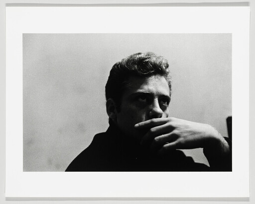 A young man sits pensive with his hand covering his mouth, staring off to the side.