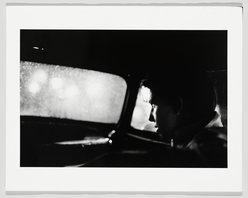 Young man sits in the backseat of a car at night, looking out a rain-speckled window.