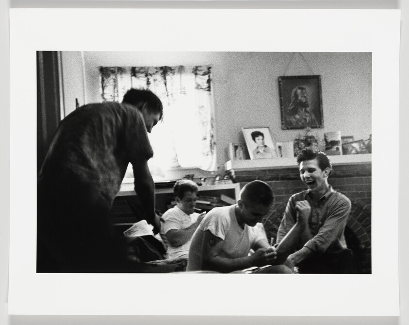 Four young men in a living room laugh and play while one tends to another's arm.