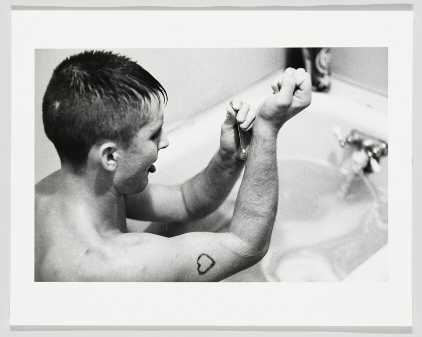 Young man in bathtub shaving arm hair with a razor, showing a small heart tattoo.