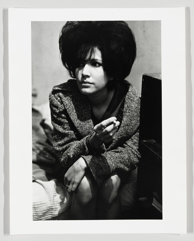 Young woman with beehive hairstyle sits on a bed holding a cigarette and looking to the side.