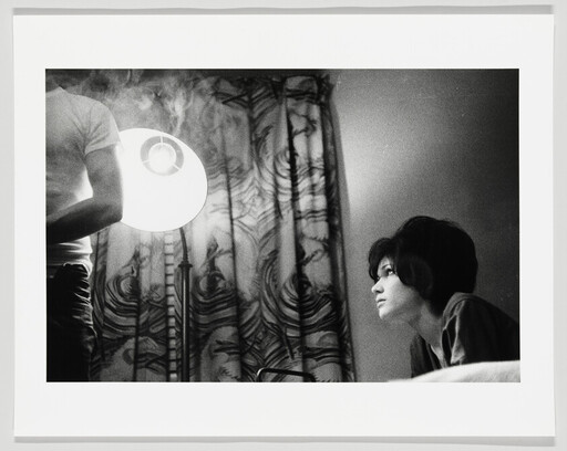 Young woman gazes toward a bright floor lamp while a standing person is partially visible.