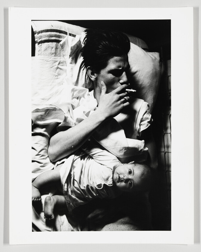 Woman lying in bed smokes a cigarette while an infant rests on her chest.