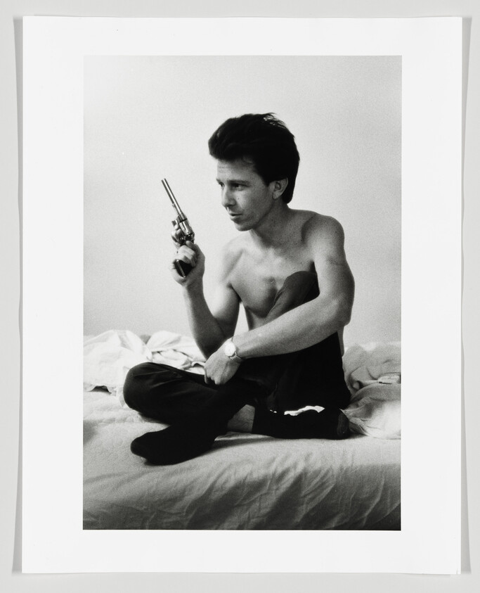 Shirtless young man sits cross-legged on a bed holding a revolver and looking aside.