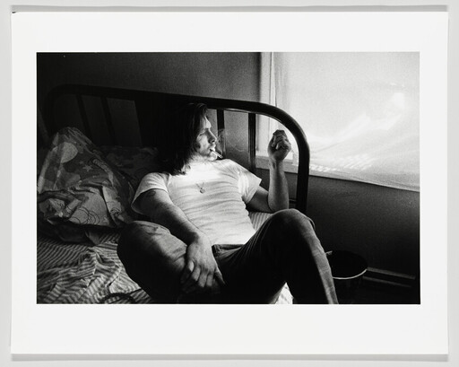 A man sits on a bed, smoking and looking out a sunlit window.