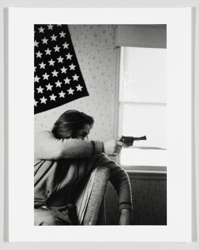 A black and white photo depicting a person sitting in a chair, resting their chin on their arm while holding a revolver. In the background, an upside-down American flag hangs on the wall next to a window with daylight coming through.