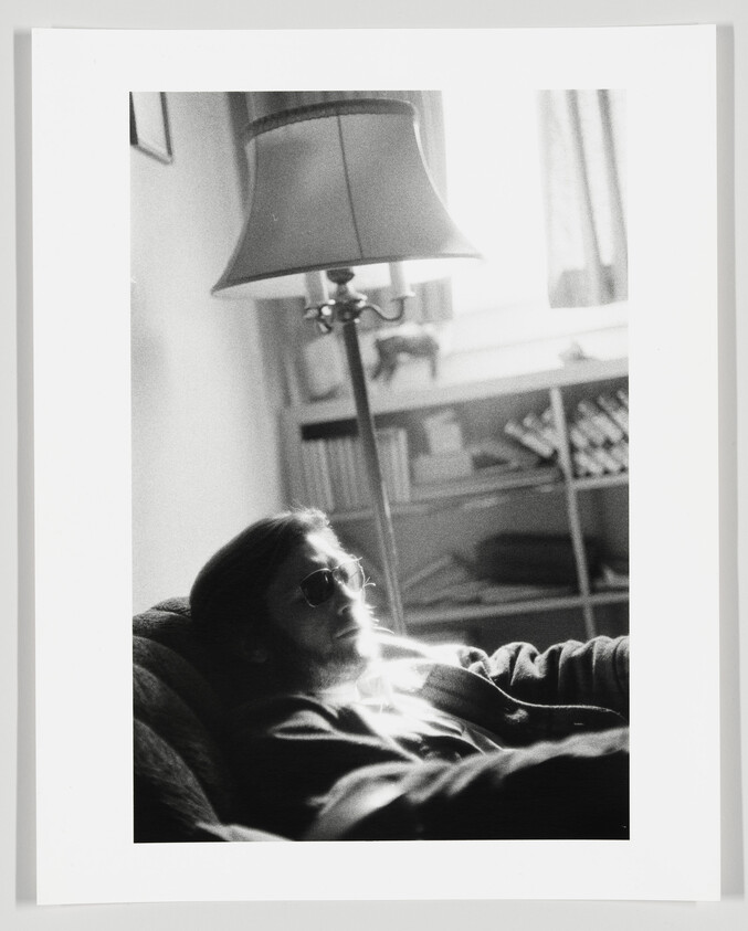 A black and white photo depicting a person lounging in a chair with sunlight streaming across their body, wearing sunglasses and a relaxed expression. A floor lamp is visible to the left, and a bookshelf in the background to the right.