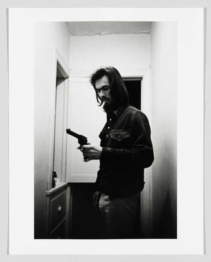 A black and white photo depicting a man standing in a doorway, looking down at a handgun he is holding in his right hand. He has a contemplative expression, long hair, and a beard, and is wearing a denim jacket and jeans. The image has a grainy texture, adding to the dramatic and intense atmosphere.