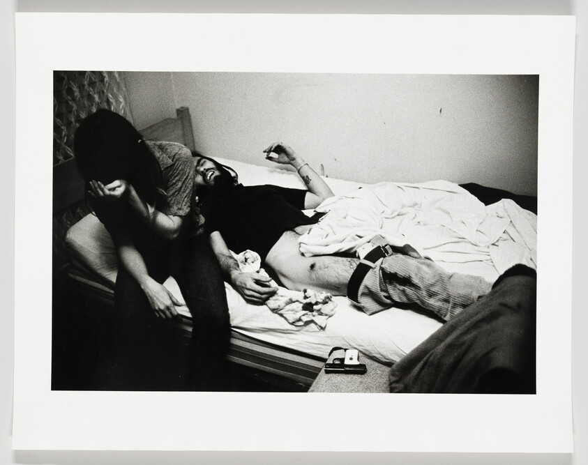 A black and white photograph capturing an emotional moment with two individuals on a bed; one person is lying down with a pained expression while the other sits beside them, covering their face with their hands, suggesting distress or sadness. There are tissues scattered on the bed, and a watch and a pager are visible on the person lying down. The setting appears to be a simple room with plain walls and a patterned curtain in the background.