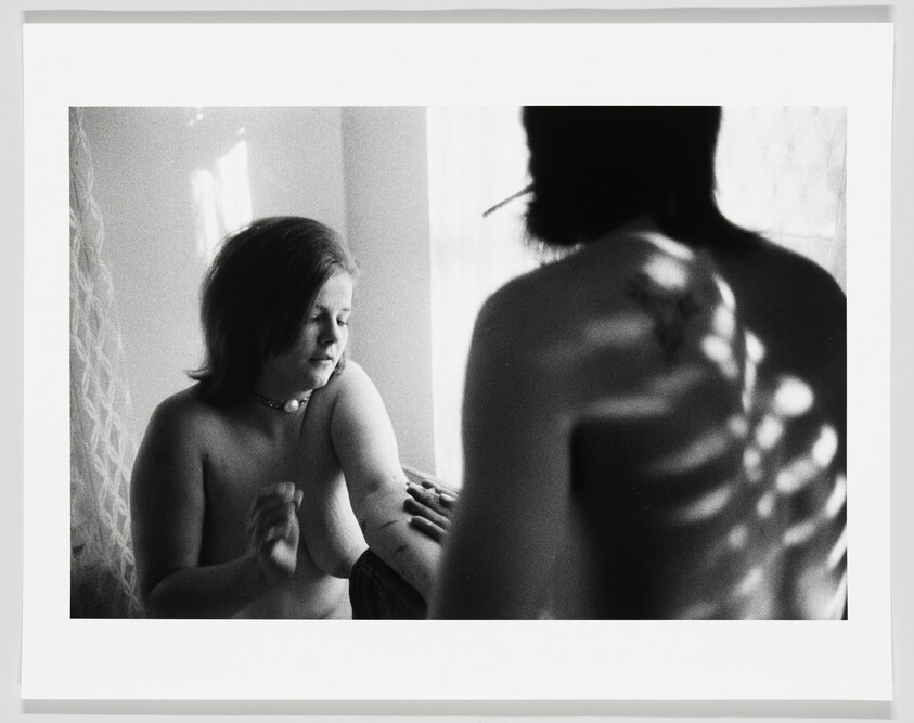 Black and white photograph capturing an intimate moment between two individuals, with a focus on the contrast of light and shadow. A woman is seated in the foreground, looking down contemplatively, while the silhouette of a person's back is visible in the background, with light casting patterns on their skin.