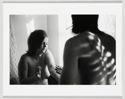 A woman examines her arm while a shirtless man with a cigarette faces away, light pattern on his back.