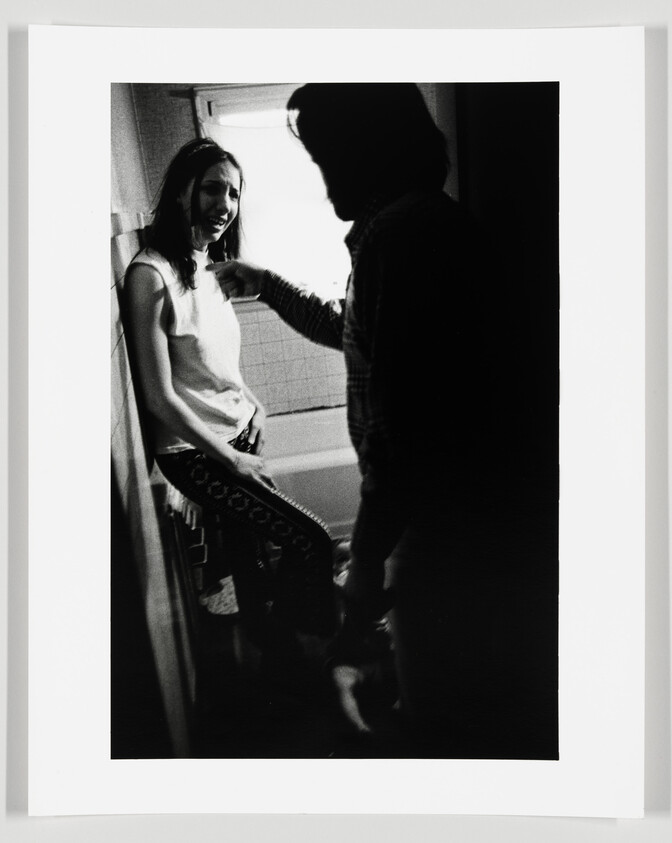 A black and white photograph capturing a candid moment between two people in a small, brightly lit room, possibly a bathroom. The woman, standing to the left, is smiling and leaning against the wall, dressed in a sleeveless top and patterned skirt. The man, facing her with his back to the camera, appears to be in mid-conversation, gesturing with his hand. The high-contrast image conveys a sense of intimacy and casual interaction.