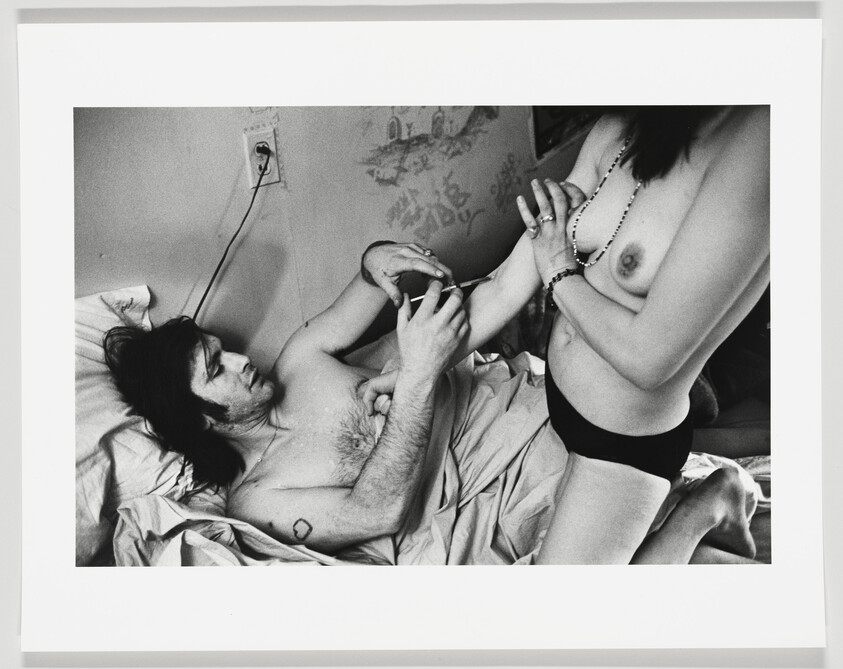 Man injects a syringe into a woman's arm while they are in a bed together.