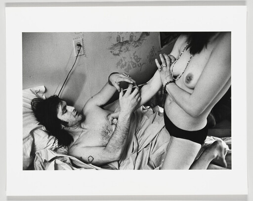 Man injects a syringe into a woman's arm while they are in a bed together.