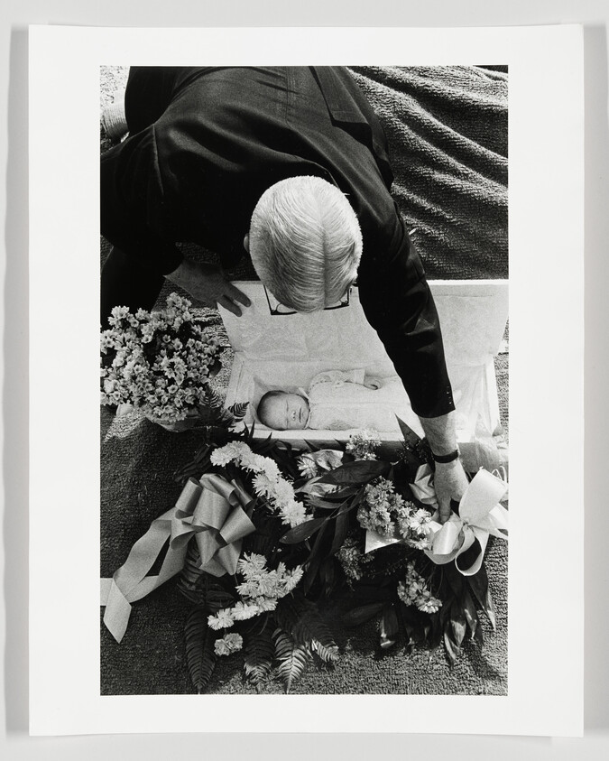 An elderly man leans over a small open casket placing a wreath beside an infant.