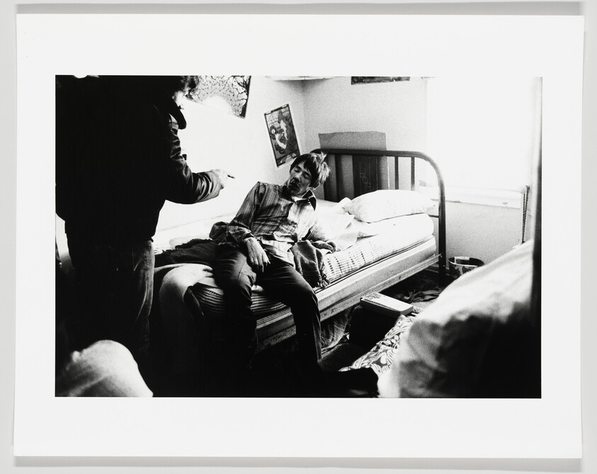 A young man sits on a bed looking toward another person who is pointing.