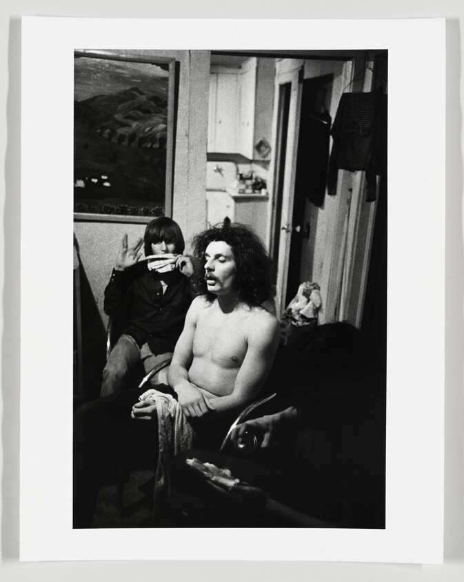 Shirtless man with curly hair sits calmly while a person behind holds a harmonica to their mouth.