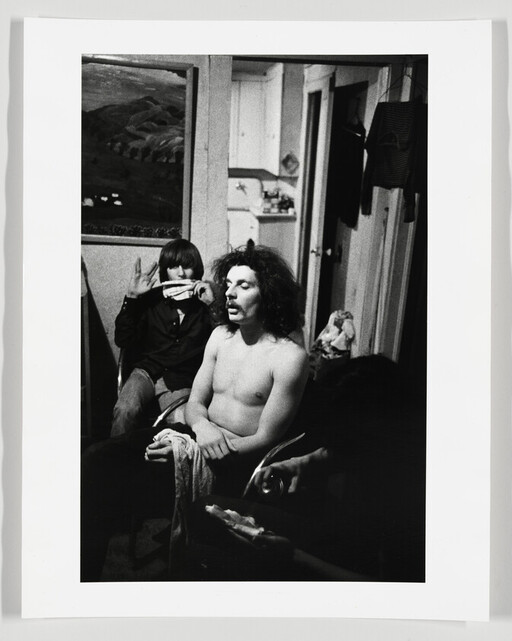 Shirtless man with curly hair sits calmly while a person behind holds a harmonica to their mouth.
