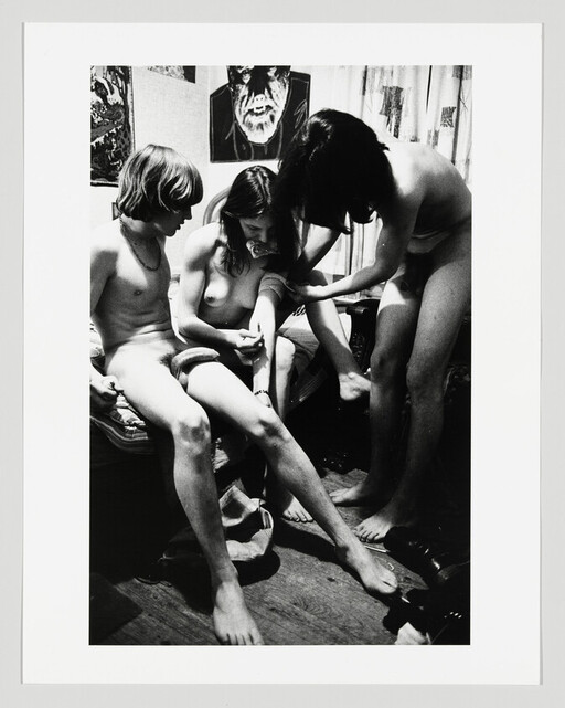Three people sit closely in a bedroom while one holds another's arm and a third watches.