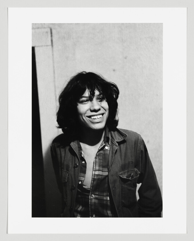 Young person with shoulder-length hair smiling while wearing a jacket and plaid shirt against a wall.