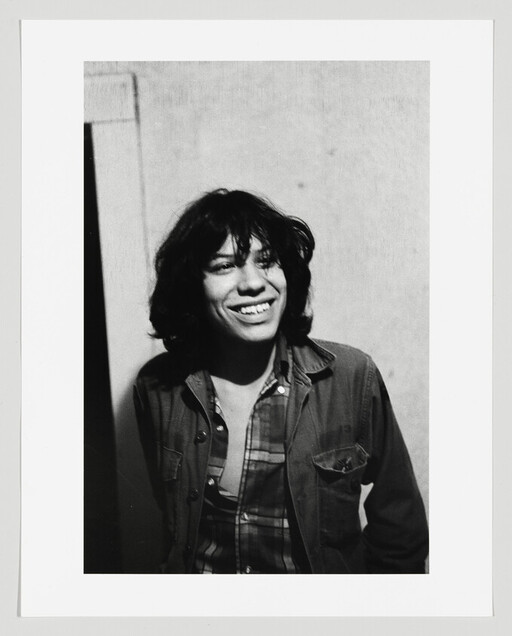 Young person with shoulder-length hair smiling while wearing a jacket and plaid shirt against a wall.