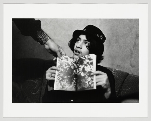 Young person in a hat holds an open book while another hand reaches to take it.