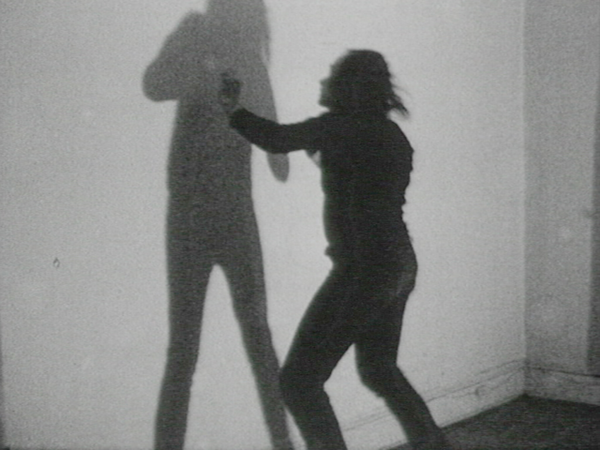 A person reaches toward and touches a tall shadow projected on a blank wall.