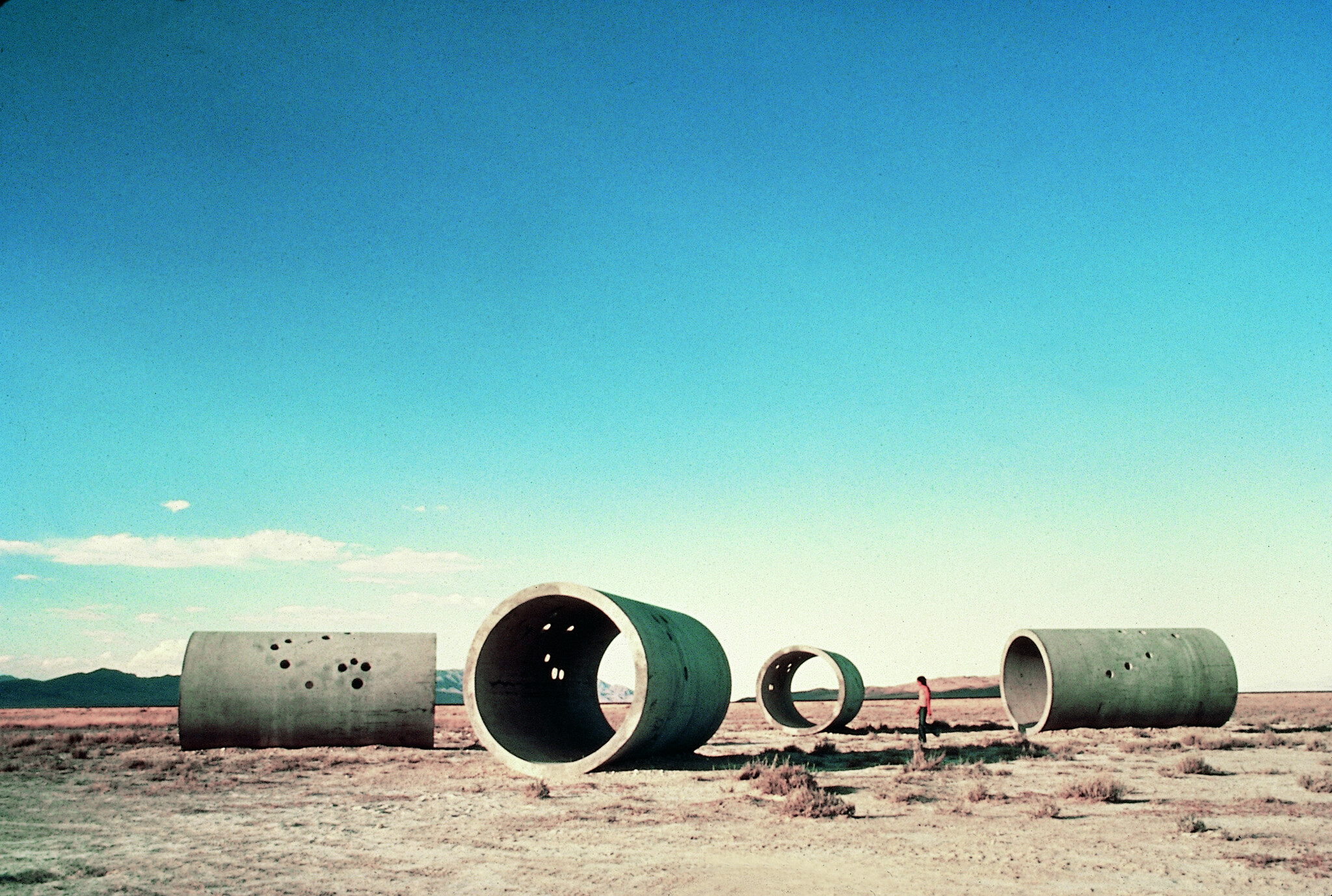 Large concrete pipe sections lie scattered in a dry field while a person walks nearby.